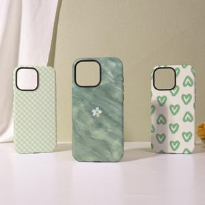 Peut inclure: Trois coques de téléphone avec des motifs différents. La première coque présente un motif vichy vert clair. La deuxième coque a un motif aquarelle vert avec une petite fleur blanche. La troisième coque a un motif de cœurs verts sur fond crème.