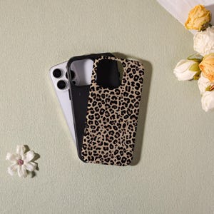 May include: A smartphone with a black case and a leopard print case. The leopard print case is tan with black spots. The smartphone is white and black. The cases are on a light green surface with flowers.
