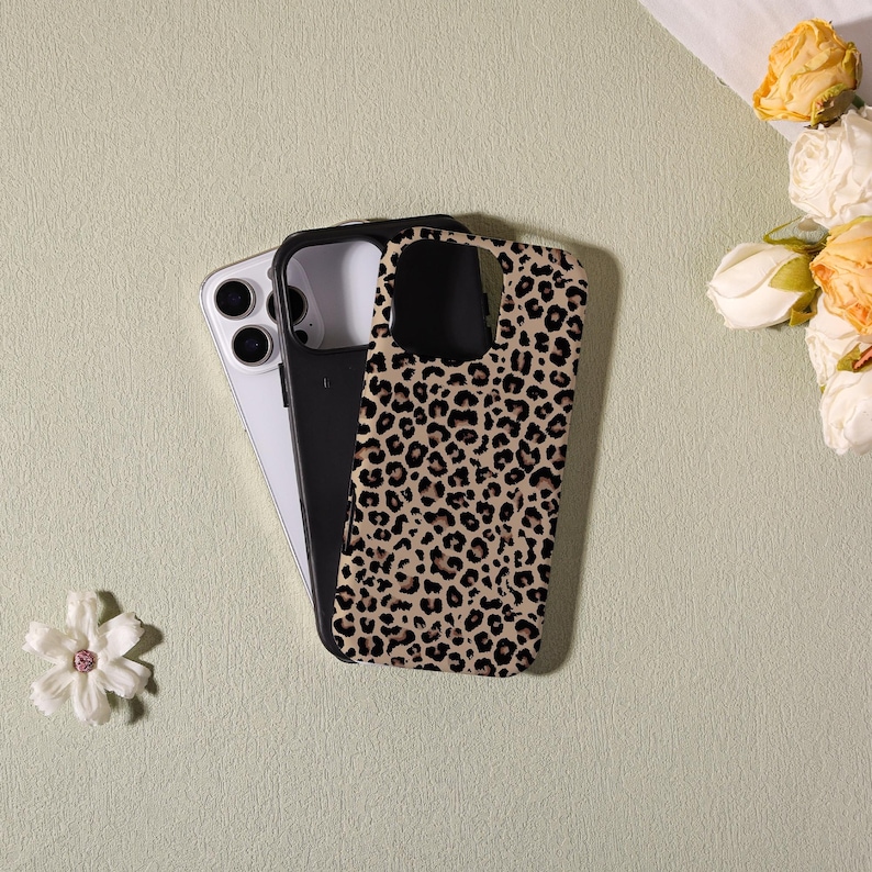 May include: A smartphone with a black case and a leopard print case. The leopard print case is tan with black spots. The smartphone is white and black. The cases are on a light green surface with flowers.
