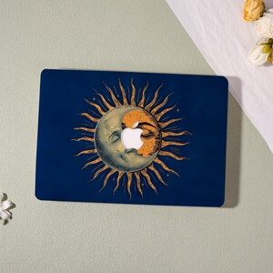 May include: A dark blue laptop skin featuring a central graphic of a crescent moon and sun face with radiating golden rays. The design incorporates a circular cutout in the center, revealing the white Apple logo underneath the celestial illustration.