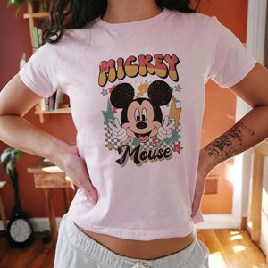 Puede incluir: Camiseta rosa claro con un dise&ntilde;o retro de Mickey Mouse. El gr&aacute;fico incluye las palabras "Mickey Mouse" en una fuente vintage colorida, con Mickey haciendo el signo de la paz. El dise&ntilde;o tiene estrellas y rayos.