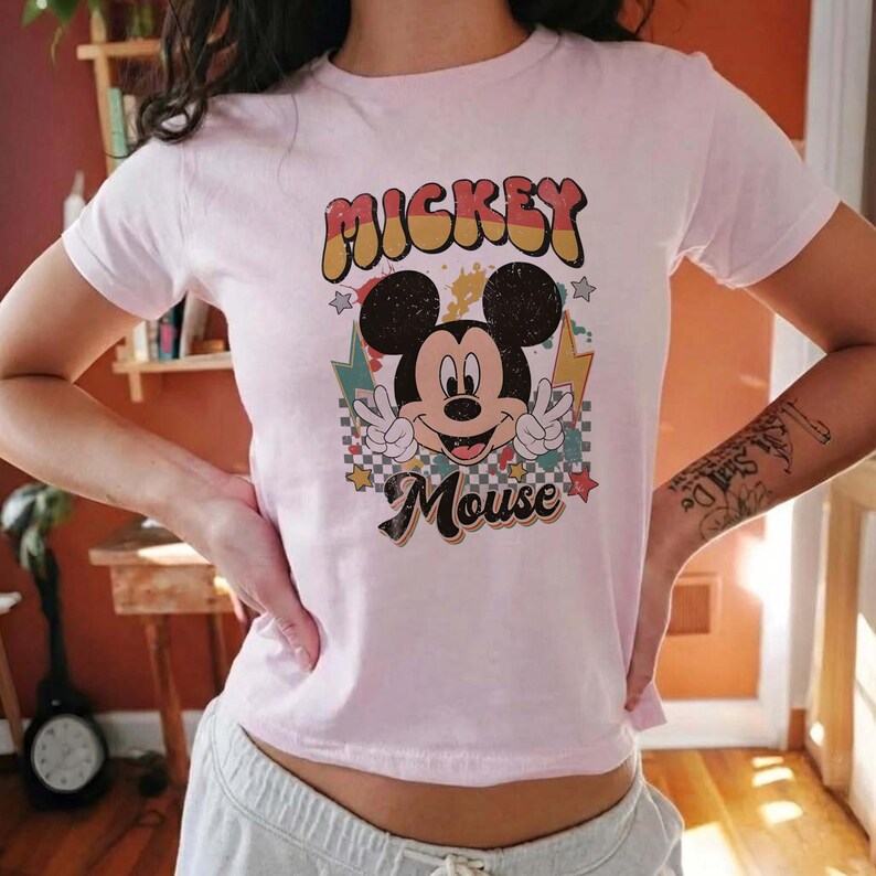 Puede incluir: Camiseta rosa claro con un dise&ntilde;o retro de Mickey Mouse. El gr&aacute;fico incluye las palabras "Mickey Mouse" en una fuente vintage colorida, con Mickey haciendo el signo de la paz. El dise&ntilde;o tiene estrellas y rayos.