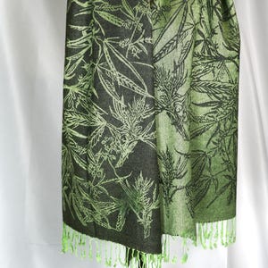 May include: A green and black shawl with a cannabis leaf pattern. The shawl has a fringe trim in a lighter green color. The design features a repeating pattern of cannabis leaves in a contrasting shade of green.