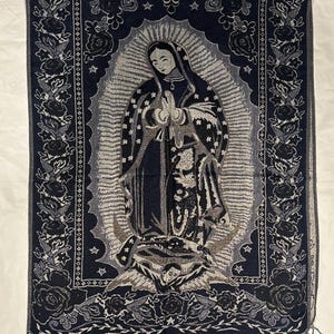 May include: A dark blue tapestry depicting the Virgin Mary with a halo, stars, and an angel. The tapestry has a floral border in shades of grey and black, and a fringed edge. The image is a religious theme.