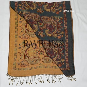 May include: A rectangular scarf featuring a dual-tone design with a mustard yellow side and a dark charcoal gray side. Both sides display a repeating paisley and floral pattern in red, green, and gold. The text RAVE MAS is printed on the fabric.