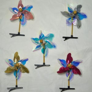 May include: Five colorful pinwheels with iridescent blades and glitter accents. Each pinwheel has a different color, including pink, silver, gold, blue, and red. They are attached to a wooden stick and a black clip.