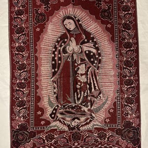 May include: A burgundy tapestry depicting the Virgin Mary in prayer. The tapestry has a floral border and a fringe at the bottom. The Virgin Mary is shown in a red robe and veil, surrounded by a halo. The tapestry is likely made of woven fabric.