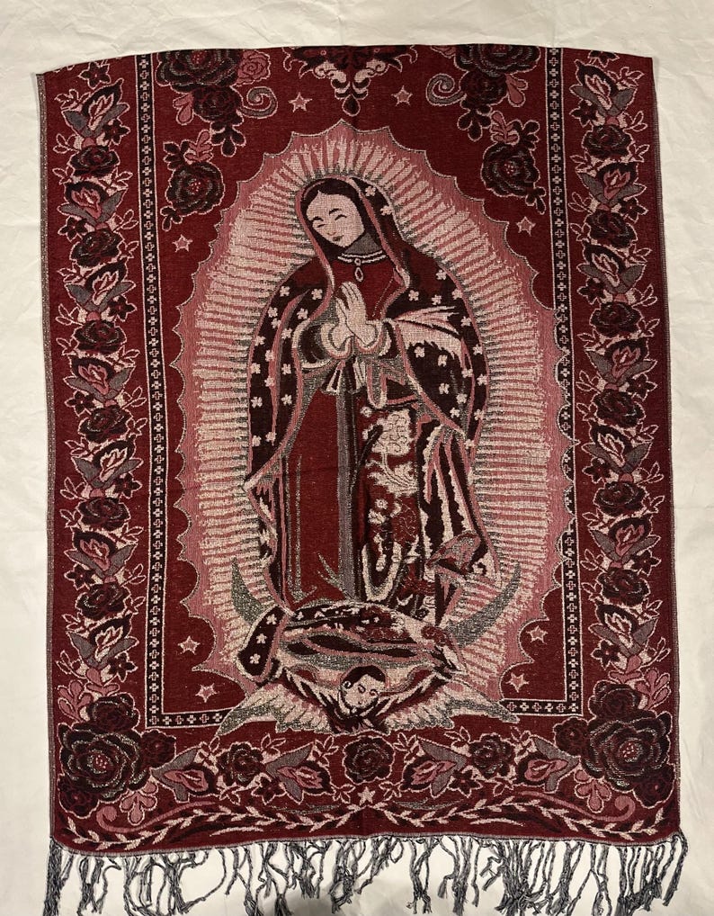 May include: A burgundy tapestry depicting the Virgin Mary in prayer. The tapestry has a floral border and a fringe at the bottom. The Virgin Mary is shown in a red robe and veil, surrounded by a halo. The tapestry is likely made of woven fabric.