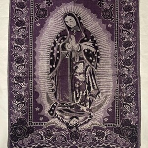 May include: A purple tapestry depicting the Virgin Mary in prayer, with a floral border of roses and a fringed edge. The image is detailed with silver and grey accents. The tapestry is likely made of woven fabric.