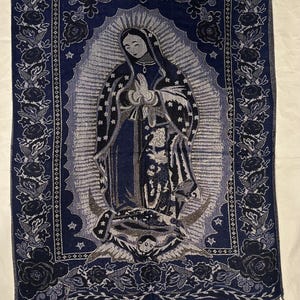 May include: A navy blue tapestry depicting the Virgin Mary in prayer. The tapestry has a floral border and fringe along the bottom. The image is in shades of blue, grey, and black. The tapestry is suitable for wall decor.
