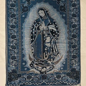 May include: Blue tapestry depicting the Virgin Mary with a halo, framed by floral and star motifs. The tapestry, with a fringed border, is mainly blue with white and grey highlights. The measurements are in centimeters.