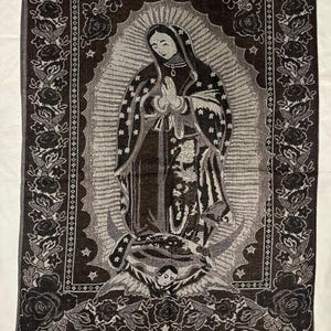 May include: A woven tapestry depicting the Virgin Mary with a halo, hands in prayer. The tapestry is mainly brown and grey, with a floral border and fringe at the base. The image is set against a white backdrop. The tapestry measures approximately 60 cm in width.