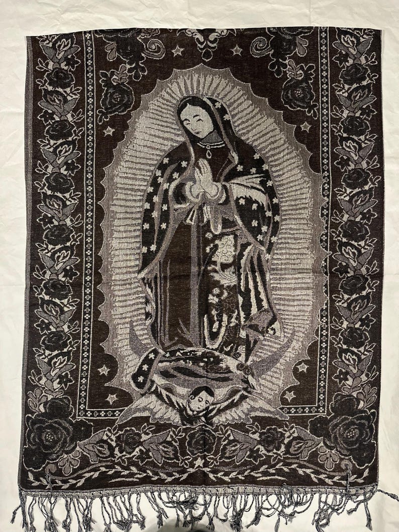 May include: A woven tapestry depicting the Virgin Mary with a halo, hands in prayer. The tapestry is mainly brown and grey, with a floral border and fringe at the base. The image is set against a white backdrop. The tapestry measures approximately 60 cm in width.