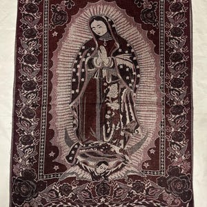May include: A burgundy tapestry featuring an image of Our Lady of Guadalupe. The tapestry has a detailed floral border and a fringed edge. The central image depicts the Virgin Mary with hands clasped in prayer.