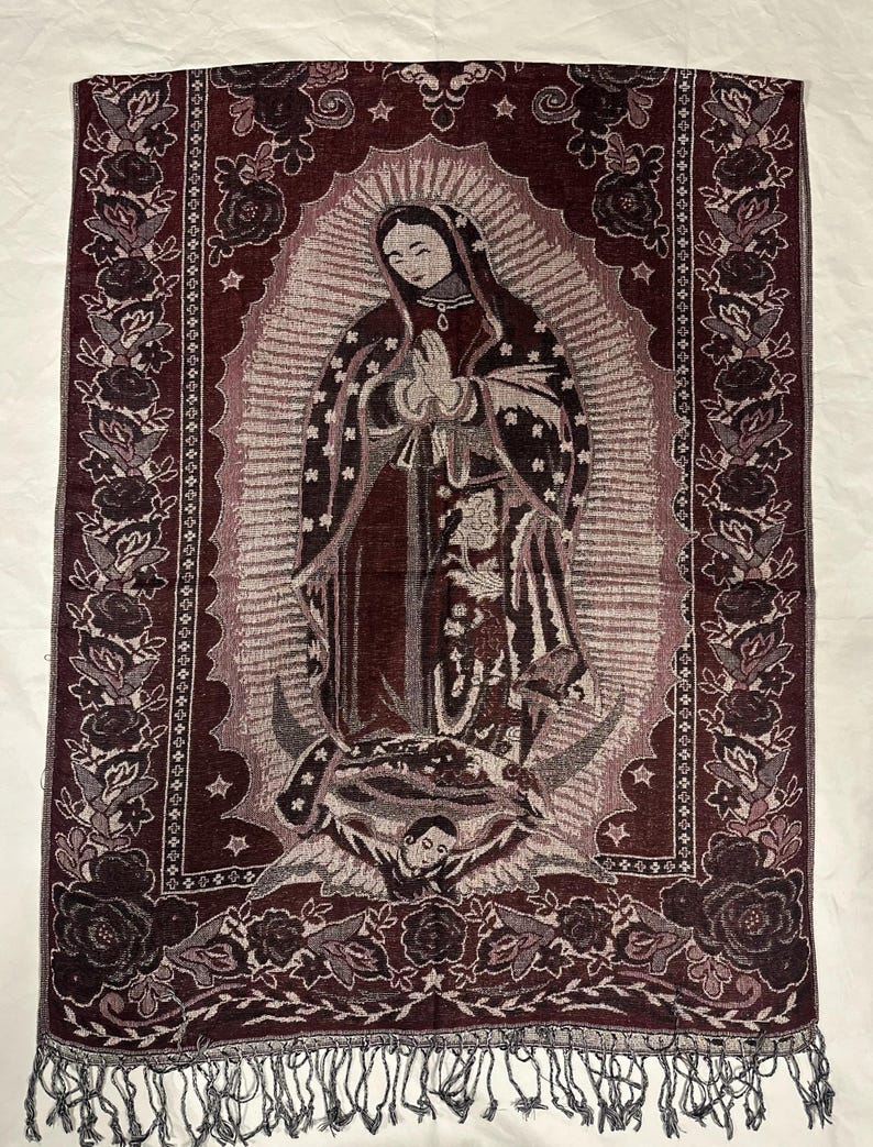 May include: A burgundy tapestry featuring an image of Our Lady of Guadalupe. The tapestry has a detailed floral border and a fringed edge. The central image depicts the Virgin Mary with hands clasped in prayer.
