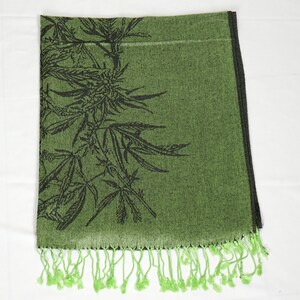 May include: Green scarf with a black cannabis leaf design. The scarf has a fringe of green tassels along the bottom edge. The scarf is folded and displayed on a white surface.
