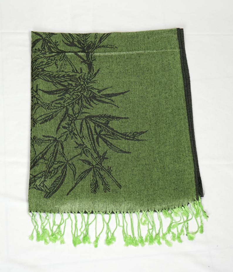 May include: Green scarf with a black cannabis leaf design. The scarf has a fringe of green tassels along the bottom edge. The scarf is folded and displayed on a white surface.