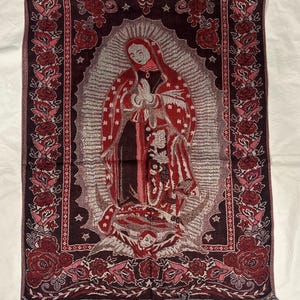 May include: A rectangular tapestry depicting the Virgin Mary in red and brown tones, framed by a floral border. The tapestry has a fringed edge and is shown on a white background.