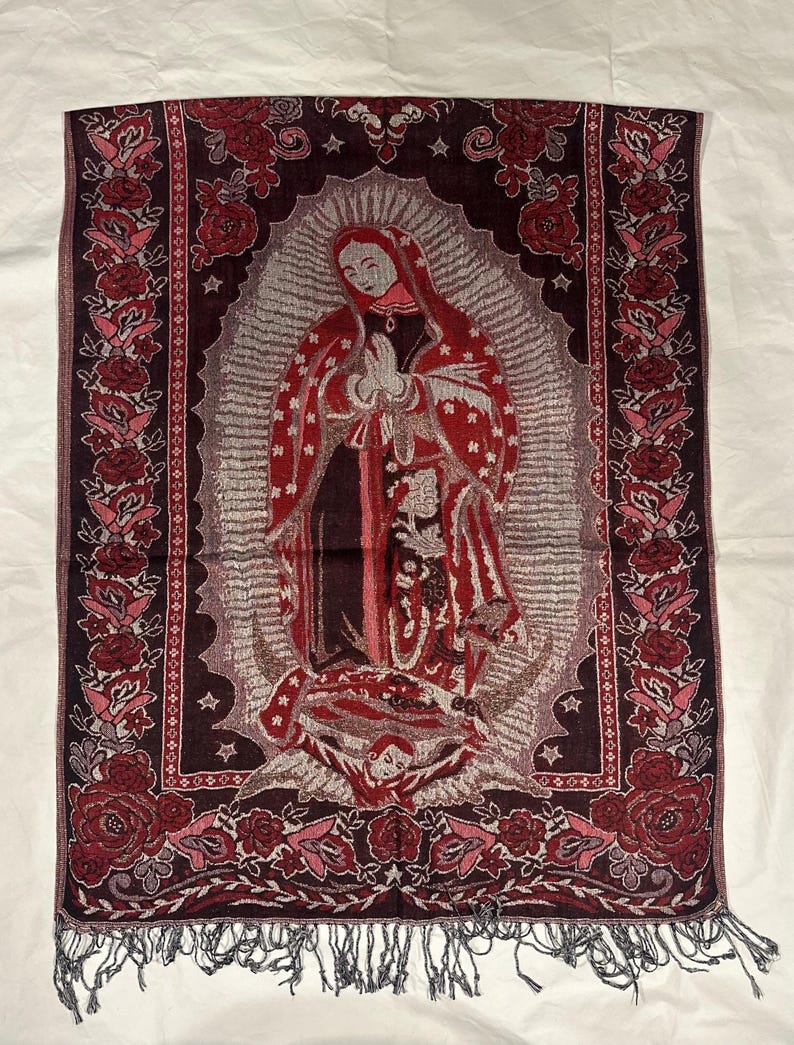 May include: A rectangular tapestry depicting the Virgin Mary in red and brown tones, framed by a floral border. The tapestry has a fringed edge and is shown on a white background.