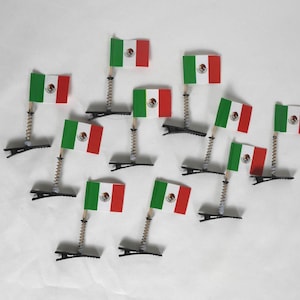 May include: A collection of small Mexican flags, each with a green, white, and red design, mounted on a spring and clip base. The flags are arranged on a white surface.