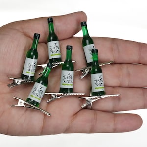May include: Miniature green soju bottle hair clips. Each clip features a small, detailed replica of a soju bottle with a white label and Korean text. The clips are attached to silver-colored metal hair clips.
