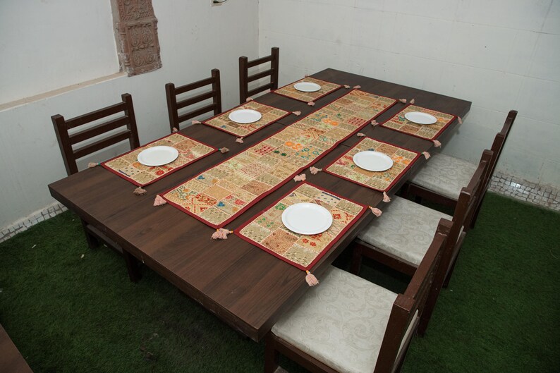 Puede incluir: Una mesa de comedor rectangular de madera marr&oacute;n oscuro con ocho sillas a juego que tienen cojines de color claro. La mesa est&aacute; decorada con un camino de mesa de patchwork largo y seis manteles individuales rectangulares a juego, cada uno con un plato blanco.