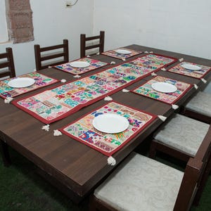 Puede incluir: Una mesa de comedor de madera marr&oacute;n oscuro con seis sillas que tienen cojines de color claro. La mesa est&aacute; decorada con un camino de mesa rectangular largo, bordado y multicolor, y seis manteles individuales a juego con un plato blanco.