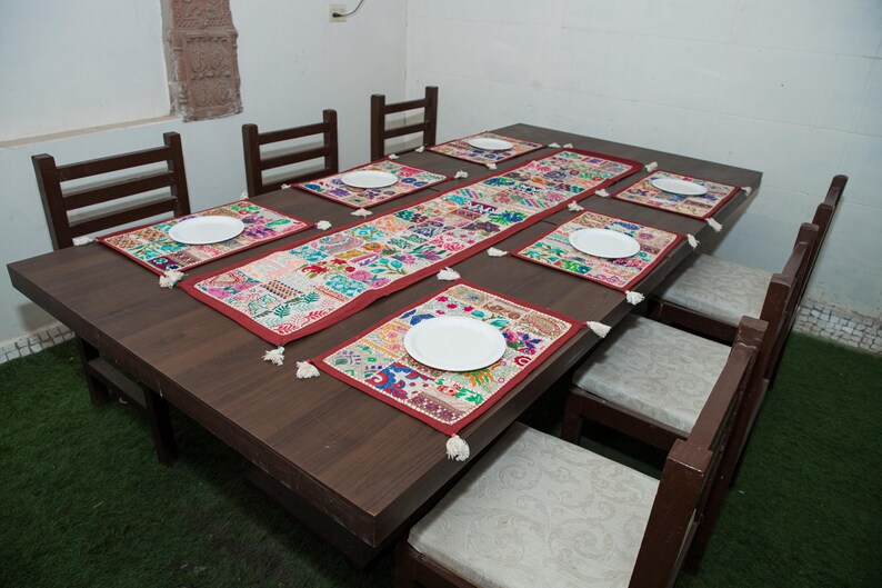 Puede incluir: Una mesa de comedor de madera marr&oacute;n oscuro con seis sillas que tienen cojines de color claro. La mesa est&aacute; decorada con un camino de mesa rectangular largo, bordado y multicolor, y seis manteles individuales a juego con un plato blanco.