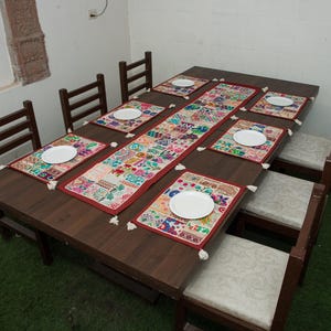 Puede incluir: Una mesa de comedor rectangular de madera marr&oacute;n oscuro con seis sillas a juego que tienen asientos tapizados de color claro. La mesa est&aacute; decorada con un camino de mesa de tela bordada multicolor y seis manteles individuales a juego, cada uno con un plato blanco.