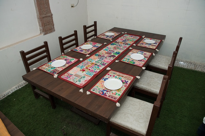 Puede incluir: Una mesa de comedor rectangular de madera marr&oacute;n oscuro con seis sillas a juego que tienen asientos tapizados de color claro. La mesa est&aacute; decorada con un camino de mesa de tela bordada multicolor y seis manteles individuales a juego, cada uno con un plato blanco.