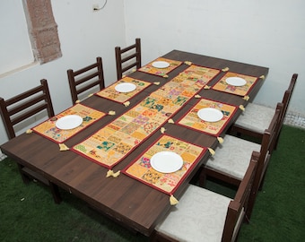 May include: A rectangular dark brown wooden dining table set with six chairs featuring light-coloured cushions. The table is decorated with a long patchwork table runner and six matching rectangular placemats, each topped with a white plate.