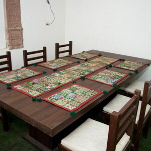 Puede incluir: Una mesa de comedor de madera marr&oacute;n oscuro con seis sillas a juego con cojines blancos. La mesa est&aacute; decorada con un camino de mesa rectangular largo y seis manteles individuales cuadrados, todos con un dise&ntilde;o de patchwork multicolor, bordes rojos y borlas verdes.