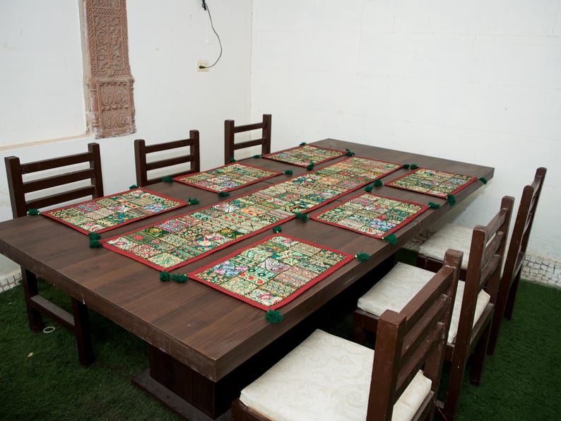 Puede incluir: Una mesa de comedor de madera marr&oacute;n oscuro con seis sillas a juego con cojines blancos. La mesa est&aacute; decorada con un camino de mesa rectangular largo y seis manteles individuales cuadrados, todos con un dise&ntilde;o de patchwork multicolor, bordes rojos y borlas verdes.