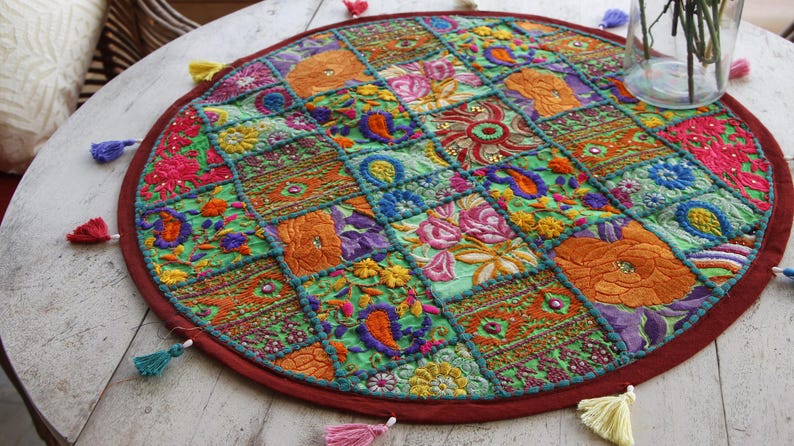 Pu&ograve; includere: Un sottopentola rotondo e colorato in patchwork con bordo rosso e nappe decorative. Il sottopentola presenta ricami floreali e paisley nei toni dell'arancione, rosa, verde e viola. &Egrave; posto su un tavolo di legno bianco.