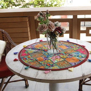 May include: A circular, white wooden table with a vibrant, patterned table mat and a vase of flowers. Two wicker chairs with red cushions and white pillows are positioned around the table. The scene is on a balcony.
