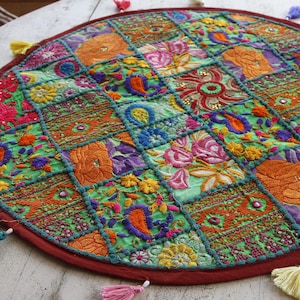 K&ouml;nnte beinhalten: Runder Tischset im Patchwork-Stil mit einem lebendigen Blumenmuster. Das Tischset zeigt eine Vielzahl von farbenfrohen, bestickten Quadraten in Gr&uuml;n-, Orange-, Rosa- und Blaut&ouml;nen. Es hat einen roten Rand und ist mit kleinen Quasten am Rand verziert.