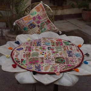 May include: A round, patchwork-style table cover featuring a variety of colourful, embroidered floral and geometric fabric squares. The circular cover is finished with a dark red border and small, multi-coloured fabric tassels along the edge.