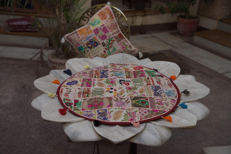 May include: A round, patchwork-style table cover featuring a variety of colourful, embroidered floral and geometric fabric squares. The circular cover is finished with a dark red border and small, multi-coloured fabric tassels along the edge.