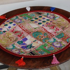May include: A round, multi-colored patchwork table mat featuring various embroidered fabric squares in shades of pink, green, blue, and gold. The mat is bordered by a solid red fabric edge and decorated with small, colorful tassels around the perimeter.