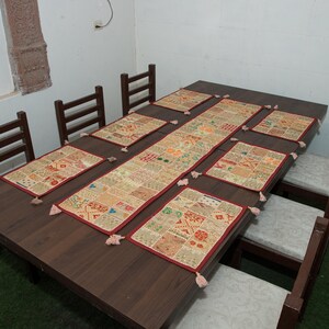 Puede incluir: Una mesa de comedor rectangular de madera marr&oacute;n oscuro con seis sillas a juego que tienen cojines de color claro. La mesa est&aacute; decorada con un camino de mesa rectangular de patchwork y seis manteles individuales rectangulares en tonos beige y rojo.