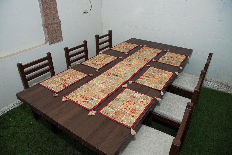 Puede incluir: Una mesa de comedor rectangular de madera marr&oacute;n oscuro con seis sillas a juego que tienen cojines de color claro. La mesa est&aacute; decorada con un camino de mesa rectangular de patchwork y seis manteles individuales rectangulares en tonos beige y rojo.