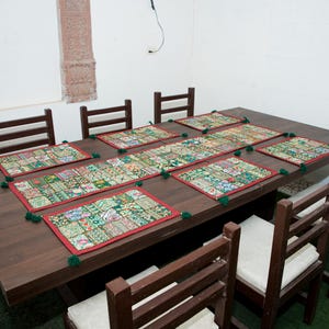 Puede incluir: Una mesa de comedor de madera marr&oacute;n oscuro con seis sillas a juego con cojines blancos. La mesa est&aacute; decorada con un camino de mesa rectangular largo y seis manteles individuales cuadrados, todos con un patr&oacute;n de patchwork colorido, bordes rojos y borlas verdes.