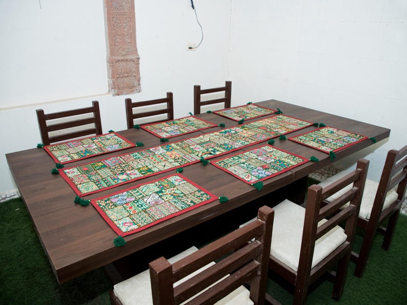 Puede incluir: Una mesa de comedor de madera marr&oacute;n oscuro con seis sillas a juego con cojines blancos. La mesa est&aacute; decorada con un camino de mesa rectangular largo y seis manteles individuales cuadrados, todos con un patr&oacute;n de patchwork colorido, bordes rojos y borlas verdes.