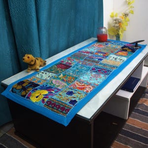 May include: A rectangular, patchwork table runner with a vibrant blue border. The runner features a variety of colorful fabric squares with floral and paisley patterns. A small, yellow dog figurine sits on the table.
