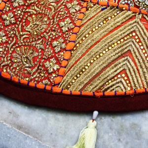 May include: Close-up of a round, decorative cushion with intricate embroidery. The cushion features a patchwork design with red, orange, and gold fabrics. Detailed gold thread embroidery and small beads add texture and visual interest. A tassel hangs from the edge.