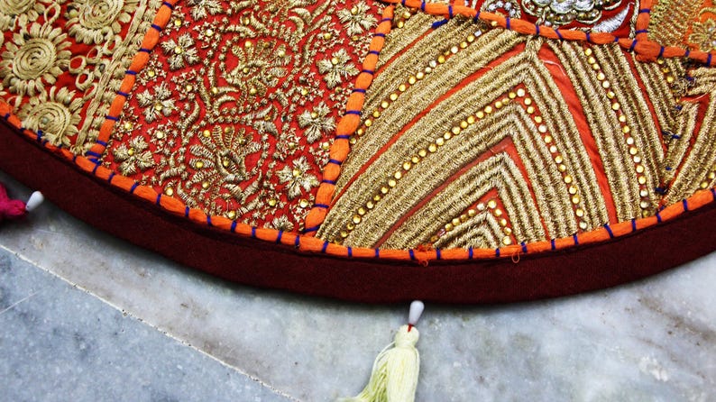 May include: Close-up of a round, decorative cushion with intricate embroidery. The cushion features a patchwork design with red, orange, and gold fabrics. Detailed gold thread embroidery and small beads add texture and visual interest. A tassel hangs from the edge.