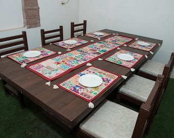May include: A dark brown wooden dining table set with six chairs featuring light-coloured cushions. The table is decorated with a long, rectangular, multicoloured embroidered table runner and six matching placemats, each topped with a white plate.