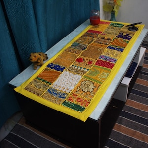 Könnte beinhalten: Ein rechteckiger Patchwork-Tischläufer mit gelbem Rand. Der Läufer weist eine Vielzahl farbiger Stoffquadrate in Gold-, Rot-, Blau- und Weißtönen auf. Ein kleiner Stoffhund sitzt auf dem Tisch neben dem Läufer.