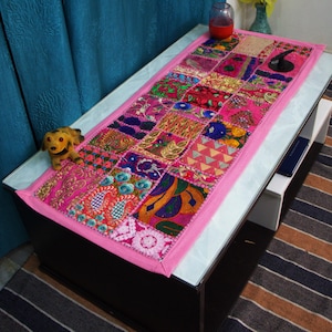 Könnte beinhalten: Rechteckiger Patchwork-Tischläufer mit rosa Rand. Der Läufer zeigt eine Vielzahl farbenfroher Stoffquadrate in Rosa, Blau, Orange und Grün. Ein kleiner Stoffhund sitzt auf dem Tisch. Der Tisch steht auf einem gestreiften Teppich.