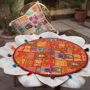 May include: A round, orange-toned fabric table mat featuring a patchwork design with intricate embroidery, metallic thread accents, and small mirrors. The mat is finished with a dark red border and several small, multi-coloured fabric tassels.